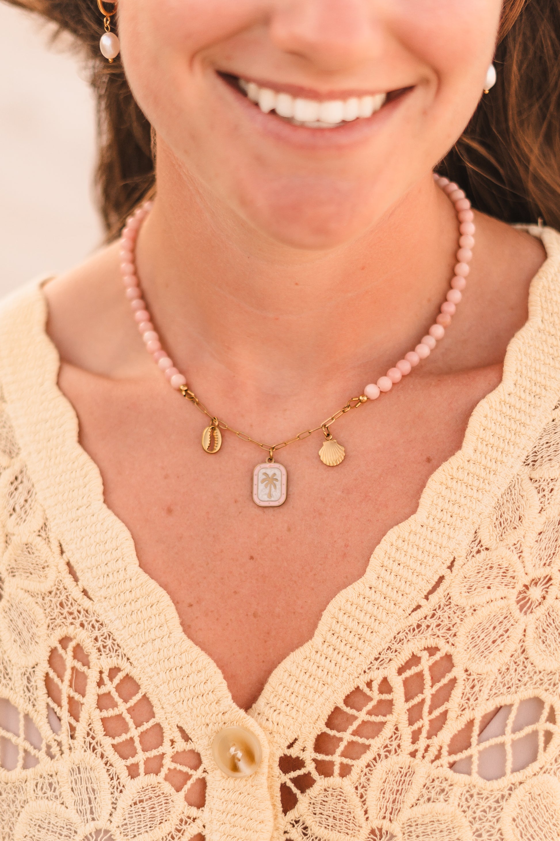 Model wearing the Malibu necklace featuring pink stabilized opal beads, a gold paperclip chain, and cowrie shell, palm tree, and scallop shell charms on waterproof 18k Gold PVD stainless steel components.