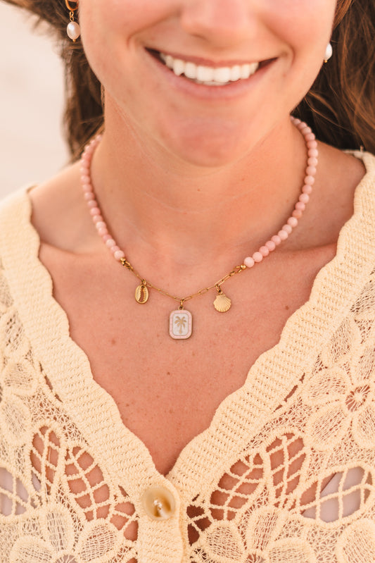 Model wearing the Malibu necklace featuring pink stabilized opal beads, a gold paperclip chain, and cowrie shell, palm tree, and scallop shell charms on waterproof 18k Gold PVD stainless steel components.