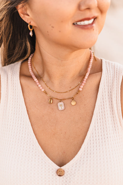 Model wearing the Malibu necklace featuring pink stabilized opal beads, a gold paperclip chain, and cowrie shell, palm tree, and scallop shell charms on waterproof 18k Gold PVD stainless steel components.